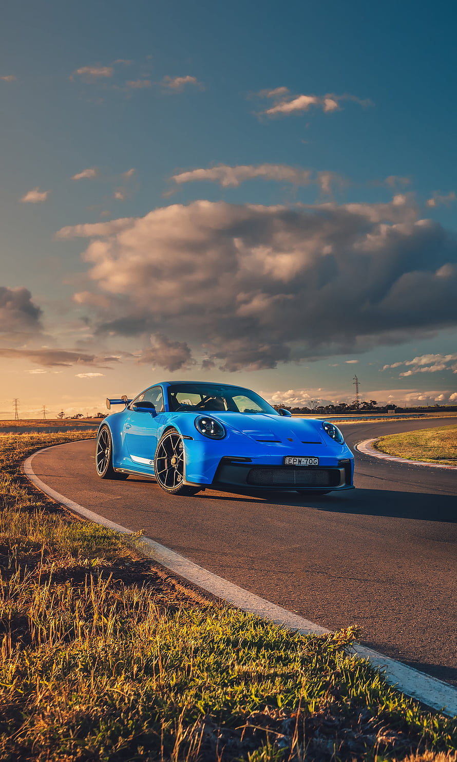 Blue Porsche 911 GT3 RS driving on a race track at sunset with dramatic clouds HD phone wallpaper