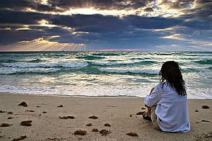 Solitary figure sitting on a sandy beach gazing at the ocean under a dramatic cloudy sky HD wallpaper