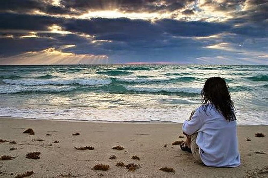 Solitary figure sitting on a sandy beach gazing at the ocean under a dramatic cloudy sky HD wallpaper