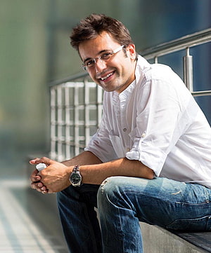 Sandeep Maheshwari smiling in white shirt and glasses sitting on a ledge HD phone wallpaper