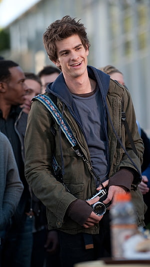 Andrew Garfield smiling with a vintage camera in an olive jacket and blue hoodie HD phone wallpaper