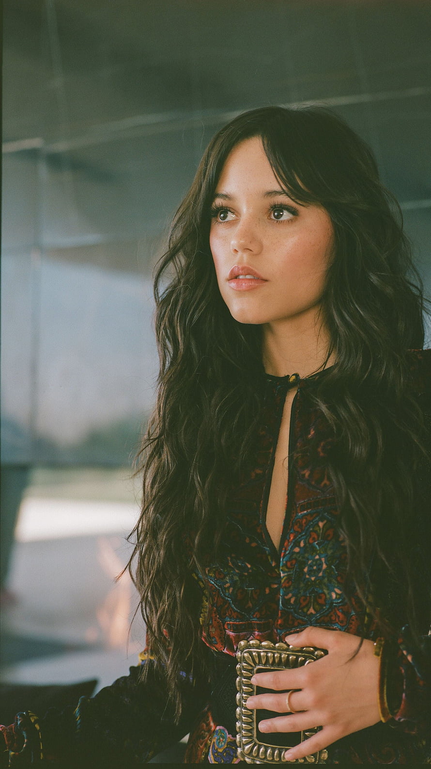 Jenna Ortega in a patterned blouse with a large gold buckle belt, gazing thoughtfully with long wavy hair HD phone wallpaper