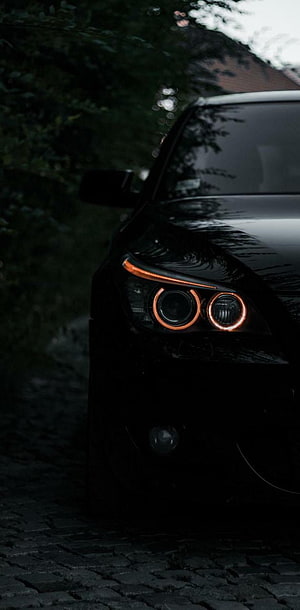 Close-up of a black BMW with glowing orange halo headlights parked on a dark cobblestone street at night HD phone wallpaper