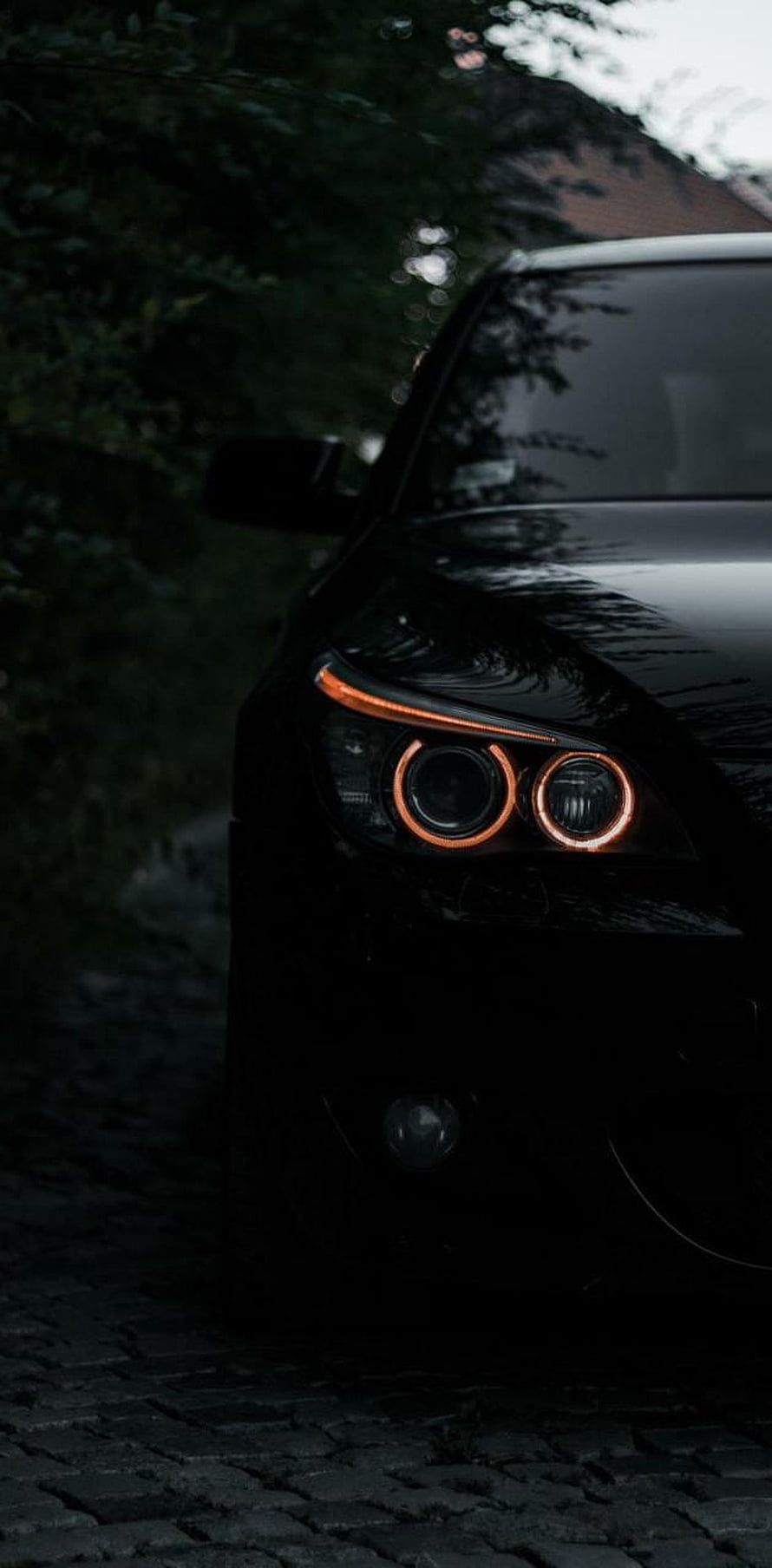 Close-up of a black BMW with glowing orange halo headlights parked on a dark cobblestone street at night HD phone wallpaper