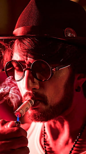 Sudeep in moody red-lit close-up wearing round sunglasses and a fedora while lighting a cigar HD phone wallpaper