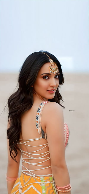 Kiara Advani in a yellow and orange patterned dress with a backless design and traditional jewelry on the beach HD phone wallpaper