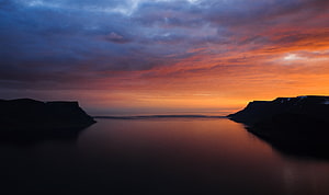 Dramatic sunset over a calm bay with silhouetted cliffs and vibrant orange sky HD wallpaper