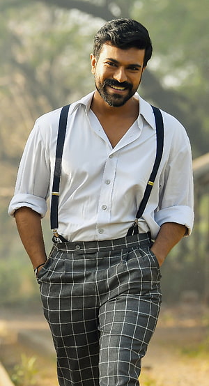 Dhanush in a white shirt and checkered trousers with suspenders smiling outdoors HD phone wallpaper