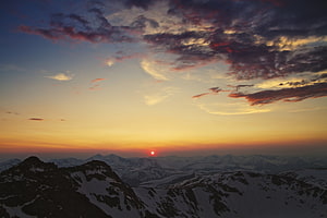 Sunset over snow-capped mountain peaks with dramatic clouds and a glowing horizon HD wallpaper