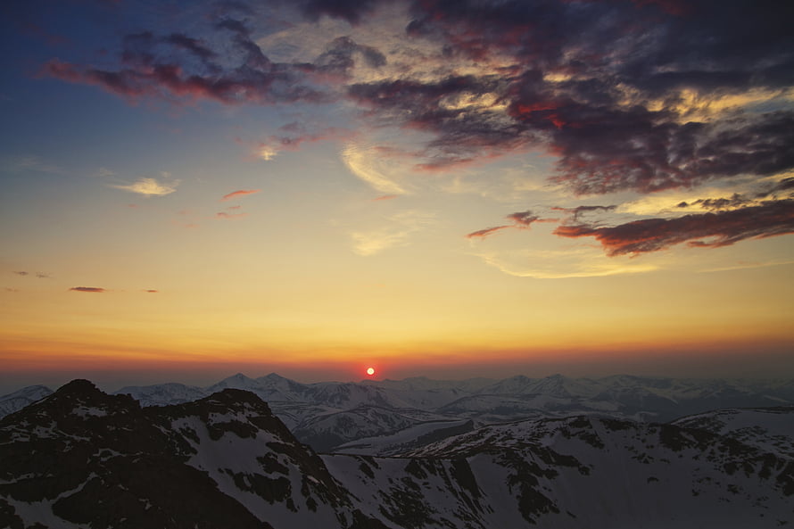 Sunset over snow-capped mountain peaks with dramatic clouds and a glowing horizon HD wallpaper