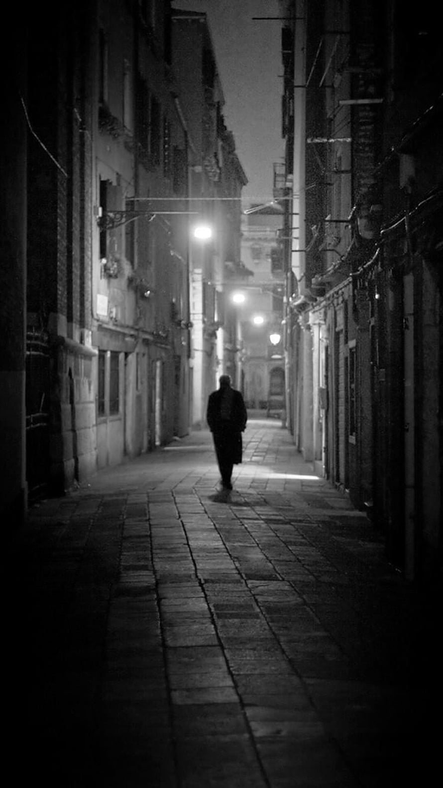 Solitary figure walking through a dimly lit cobblestone alley at night in black and white HD phone wallpaper