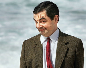 Rowan Atkinson as Mr Bean in a brown tweed jacket and red tie against a blurred ocean backdrop HD wallpaper