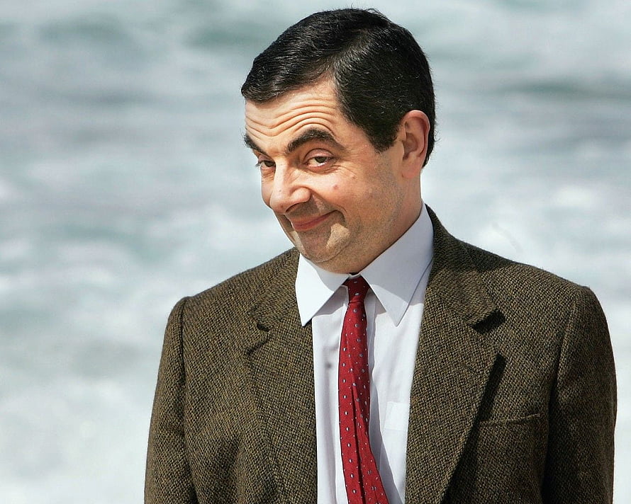 Rowan Atkinson as Mr Bean in a brown tweed jacket and red tie against a blurred ocean backdrop HD wallpaper