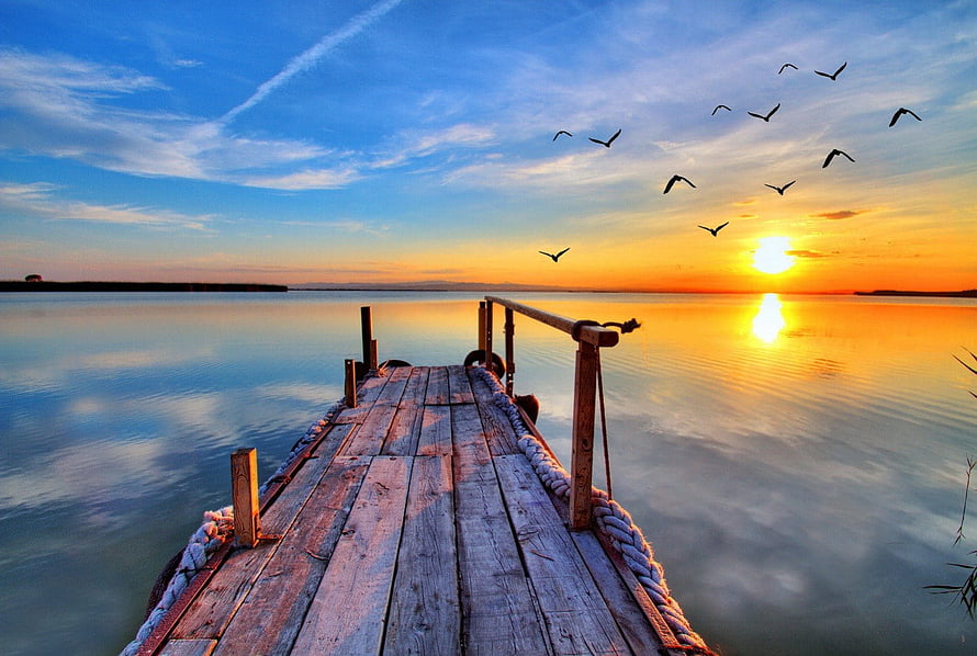 Wooden pier extending into calm lake at sunset with birds flying across golden sky HD wallpaper