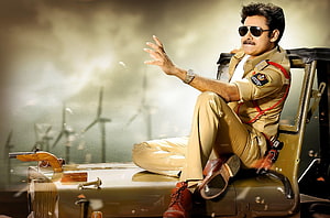 Pawan Kalyan in khaki police uniform seated on a vintage car with wind turbines in the background HD wallpaper