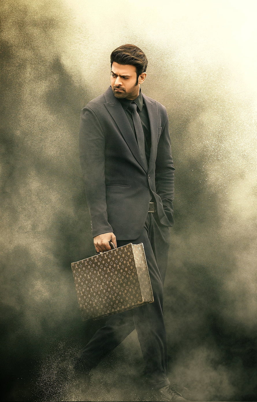 Prabhas in a dark suit holding a briefcase walking through smoke HD phone wallpaper