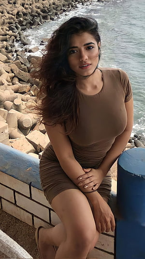 Saiyami Kher in a brown dress sitting by the seaside with ocean waves and rocks in the background HD phone wallpaper