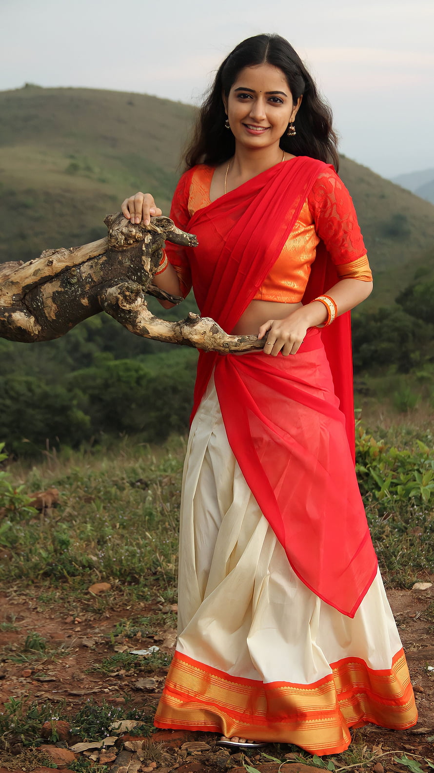 Ashika Ranganath in a vibrant red and cream saree posing with a rustic wooden prop in a scenic hilly landscape HD phone wallpaper