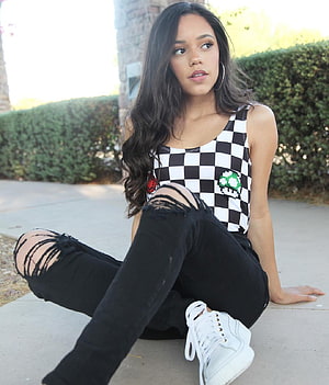 Jenna Ortega in a checkered tank top and distressed black jeans sitting outdoors HD phone wallpaper