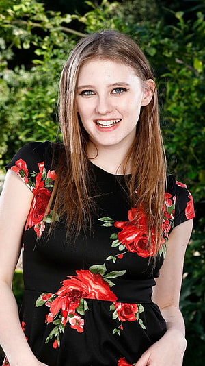 Melody Marks smiling portrait in a black dress with red rose floral print against a green garden backdrop HD phone wallpaper