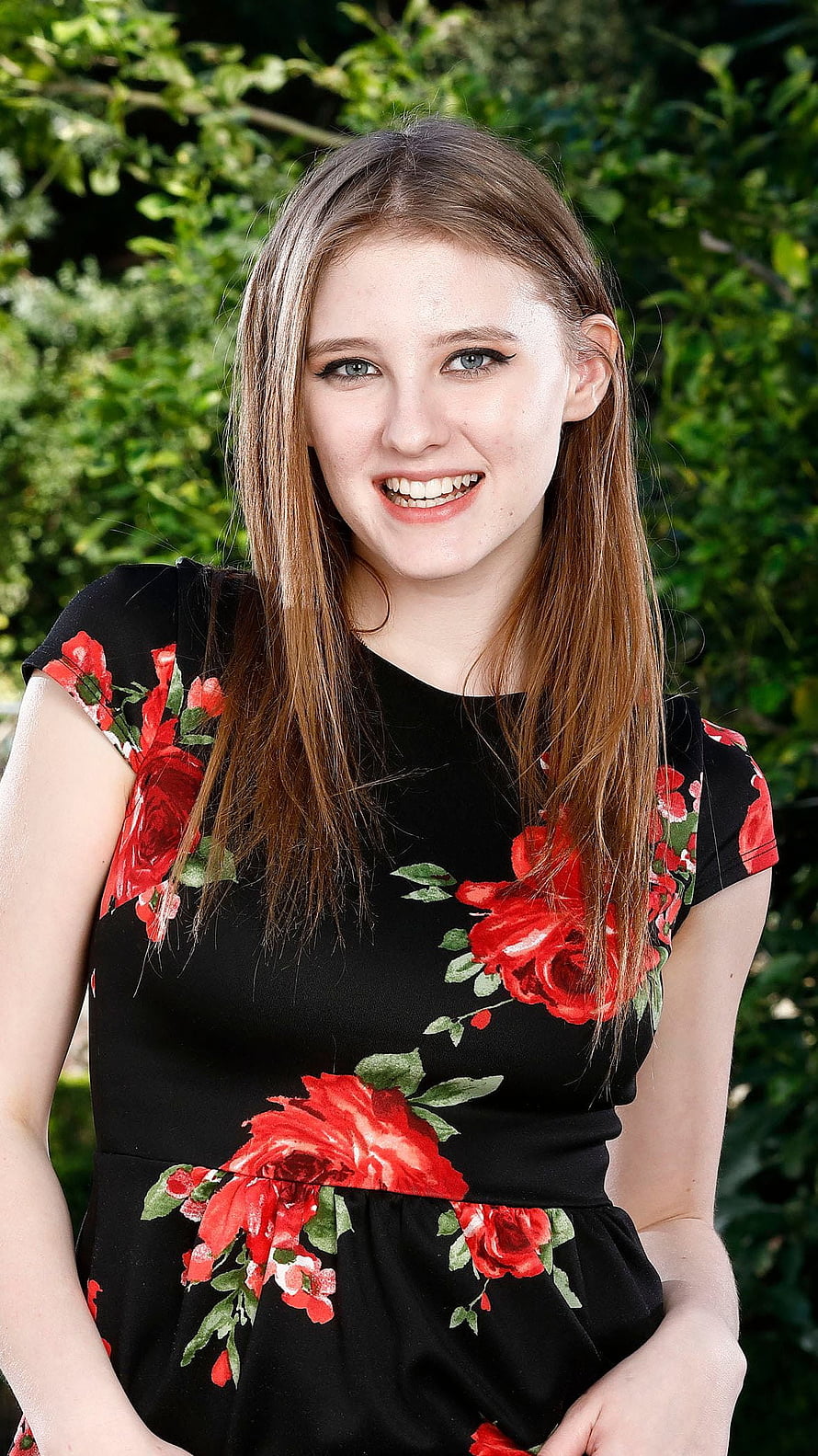 Melody Marks smiling portrait in a black dress with red rose floral print against a green garden backdrop HD phone wallpaper