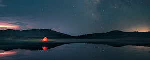 Glowing red tent on a still lake under a star-filled night sky with distant hills HD wallpaper