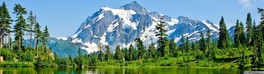 Majestic snow-capped mountain peak rising above a lush green forest and calm lake under a clear blue sky HD wallpaper
