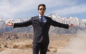 Tony Stark in a dark pinstripe suit standing with arms outstretched in a mountainous desert landscape HD wallpaper