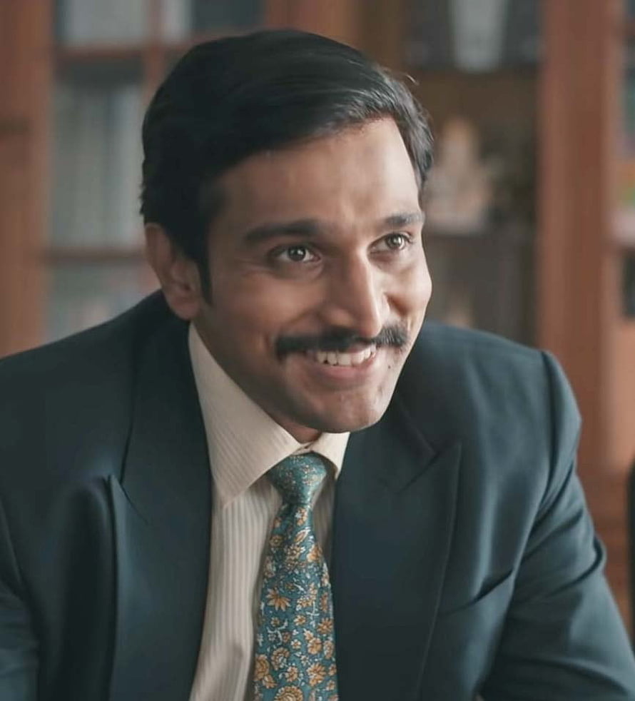 Pratik Gandhi in a dark suit and patterned tie with a warm smile in an office setting HD phone wallpaper