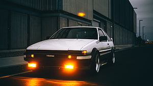 White Toyota AE86 Trueno with glowing amber headlights parked on a dark urban street at dusk HD wallpaper