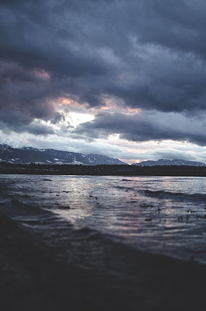 Moody Pacific Northwest shoreline with snow-capped mountains under a dramatic stormy sky HD phone wallpaper