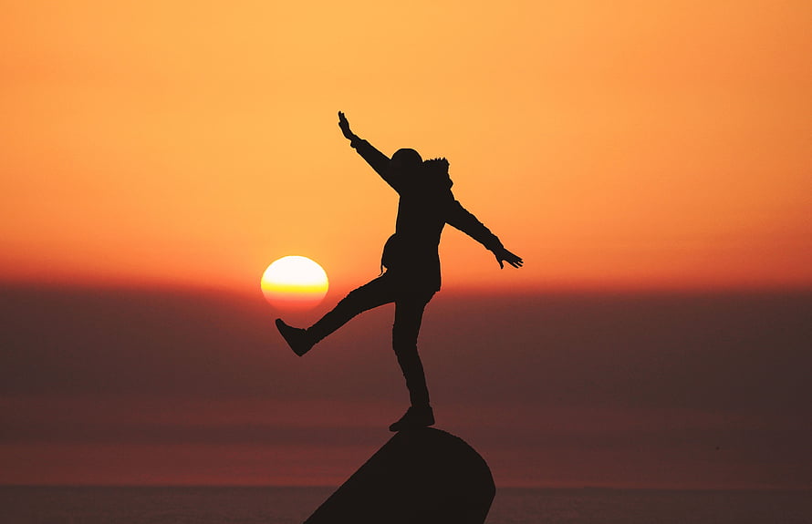 Silhouette of a person balancing on a rock against a vibrant orange sunset sky HD wallpaper
