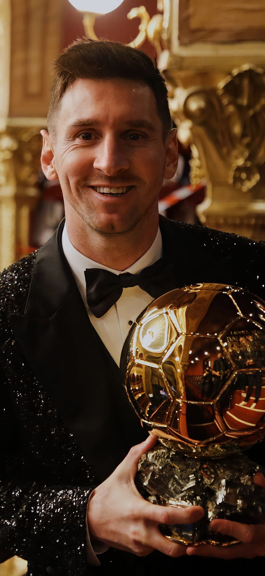 Lionel Messi in a sequined tuxedo holding the golden Ballon d'Or trophy at a formal gala HD phone wallpaper