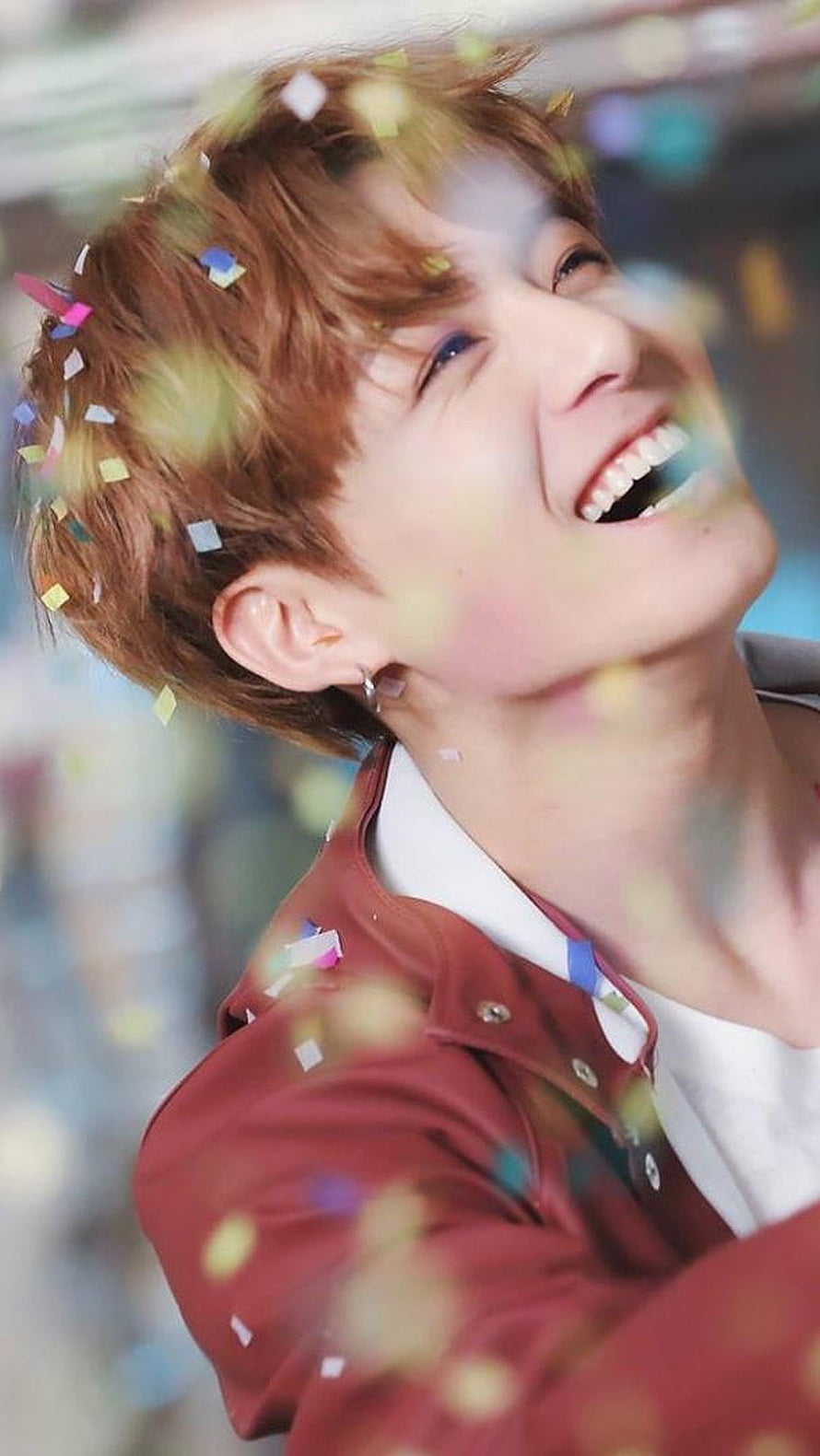 Joyful K-pop idol with brown hair laughing under colorful confetti in a red jacket HD phone wallpaper