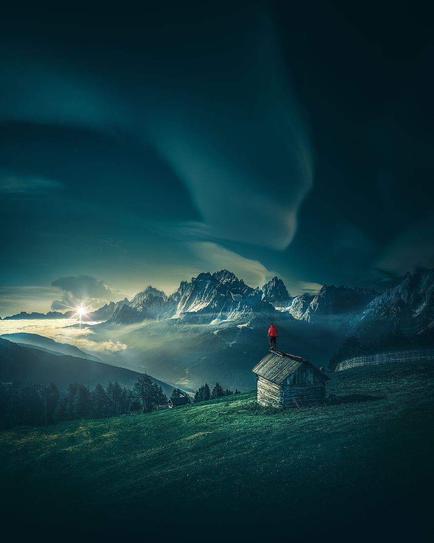 Lone Hiker on Alpine Cabin Roof at Sunrise with Dramatic Mountain Peaks and Clouds HD phone wallpaper