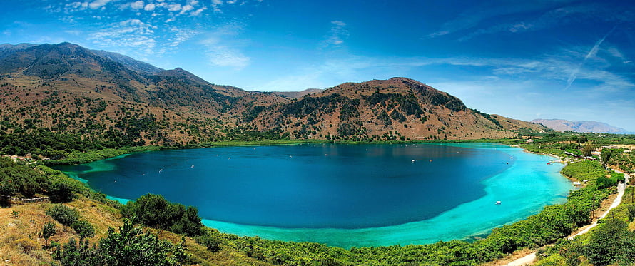 Stunning panoramic view of a turquoise lake surrounded by arid mountains and lush green vegetation under a bright blue sky HD wallpaper