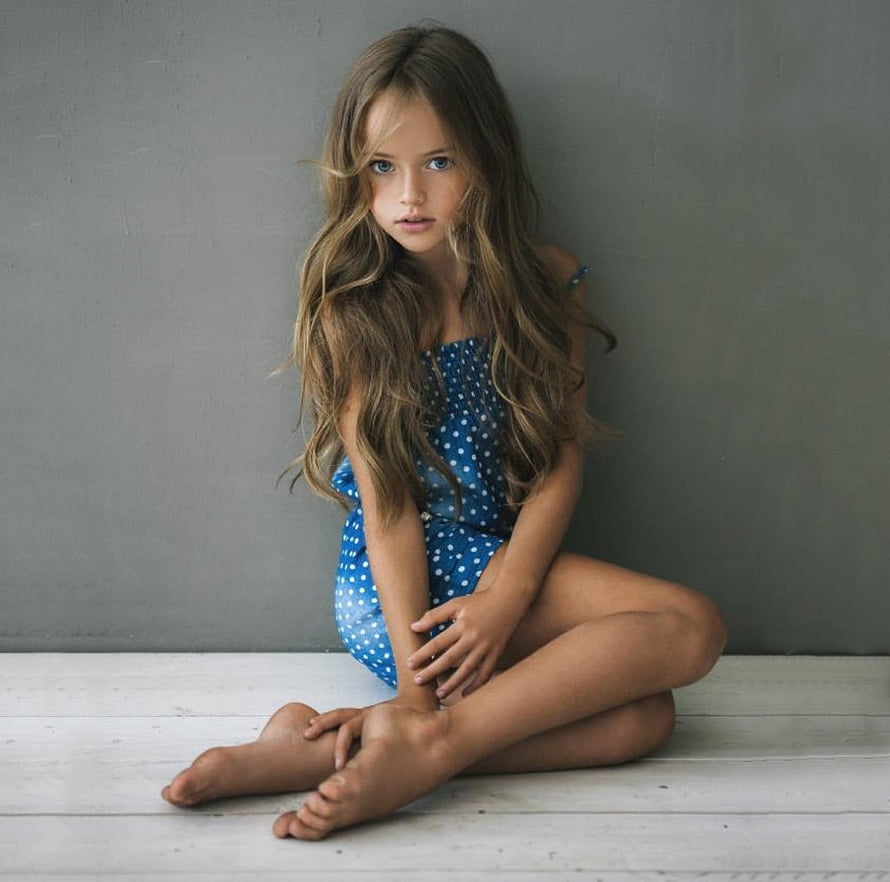Young girl with long wavy hair sitting barefoot in a blue polka dot dress against a gray wall HD wallpaper