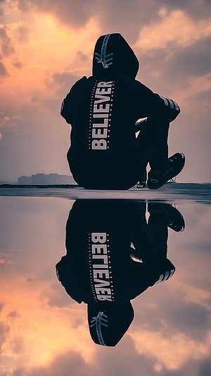 Silhouette of a person in a hoodie with Believer text sitting by water at sunset HD phone wallpaper