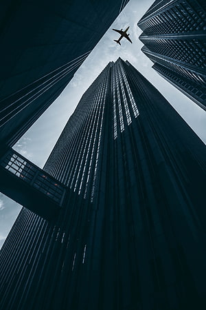 Low-angle view of towering skyscrapers with an airplane flying overhead against a cloudy sky HD phone wallpaper