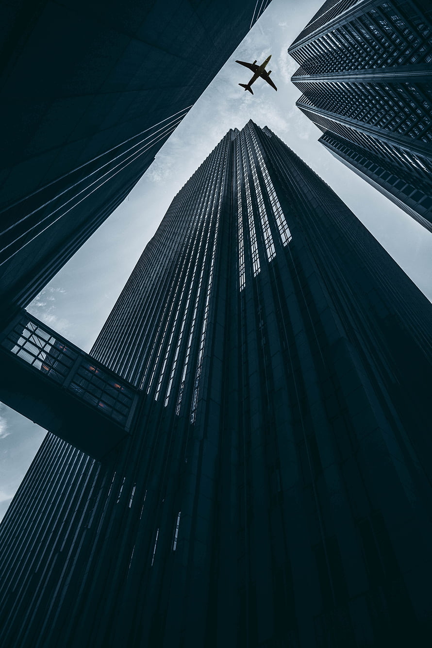 Low-angle view of towering skyscrapers with an airplane flying overhead against a cloudy sky HD phone wallpaper