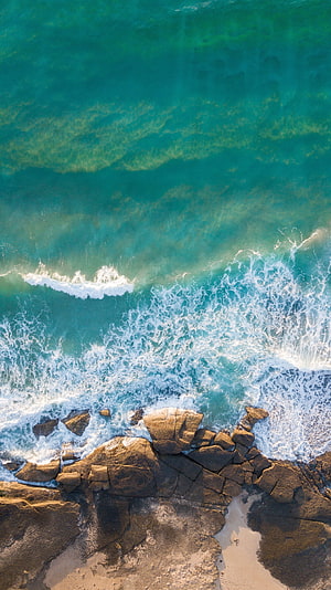 Aerial View of Turquoise Ocean Waves Crashing Against Rocky Coastline HD phone wallpaper