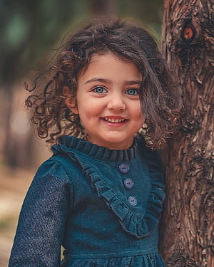 Portrait of a smiling little girl with curly hair and blue eyes wearing a teal dress leaning against a tree HD phone wallpaper