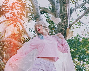 Taylor Swift in a pink crop top and light jeans posing under a tree with hanging heart ornaments and warm sunlight HD wallpaper