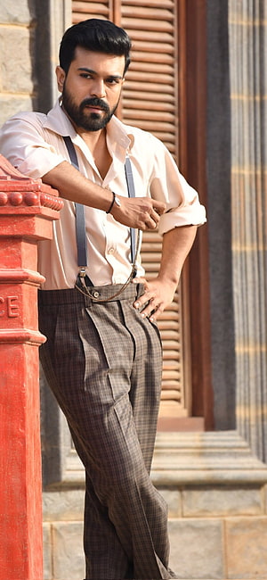 Dhanush in vintage style with suspenders and checkered trousers leaning against a red pillar HD phone wallpaper