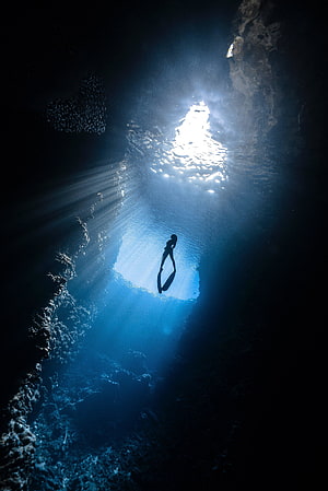 Silhouetted Free Diver Descending Through Sunlit Underwater Cavern HD phone wallpaper