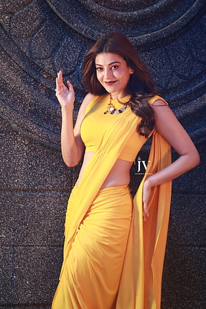 Kajal Agarwal in a vibrant yellow saree posing against a carved stone wall HD phone wallpaper