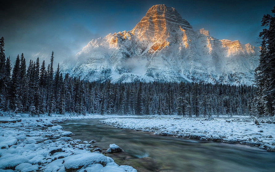 Snow-Capped Mountain Peak Rising Above a Frozen River and Evergreen Forest at Dawn HD wallpaper