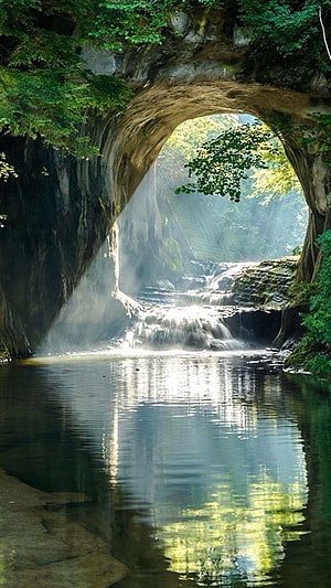 Serene waterfall cascading through a natural rock arch into a calm pool HD phone wallpaper