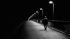 Solitary figure walking along a curved bridge at night under a row of street lamps in black and white HD wallpaper